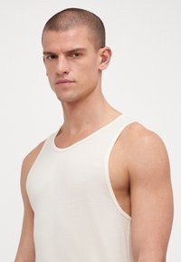Lys beige tank top med teksturstoff, bred utringning og tynne stropper. Den har kontrastfarge langs kantene og skuldersømmene.