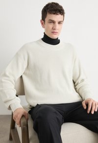 Maglione a coste color crema con scollo rotondo. Indossato sopra un dolcevita nero, con maniche ampie e una vestibilità rilassata.