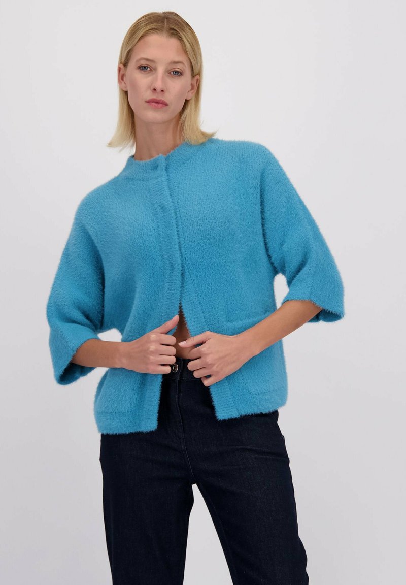Cardigan à manches courtes en bleu vif, fabriqué dans un matériau texturé avec un devant ouvert et deux poches avant, porté avec un jean foncé.