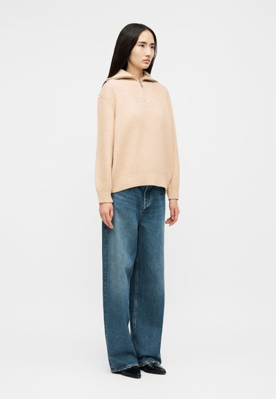 Femme debout en pull beige côtelé avec fermeture éclair à l'avant et jean bleu à jambes larges, portant des chaussures noires pointues sur fond blanc.