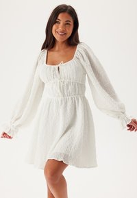 Vit, texturerad klänning med långa puffärmar, fyrkantig halsringning och dragsko i midjan som skapar en utsvängd silhuett.