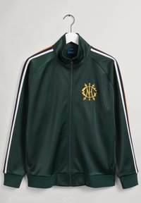 Chaqueta de chándal verde con cremallera, con rayas blancas, marinas y naranjas en las mangas. Presenta puños acanalados, un cuello alto y un logo amarillo bordado.