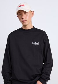 Čierna košeľa s dlhým rukávom a bielym logom "Carhartt" na hrudi. Osoba má na hlave krémovú šiltovku s červeným nápisom "Recap Racing."