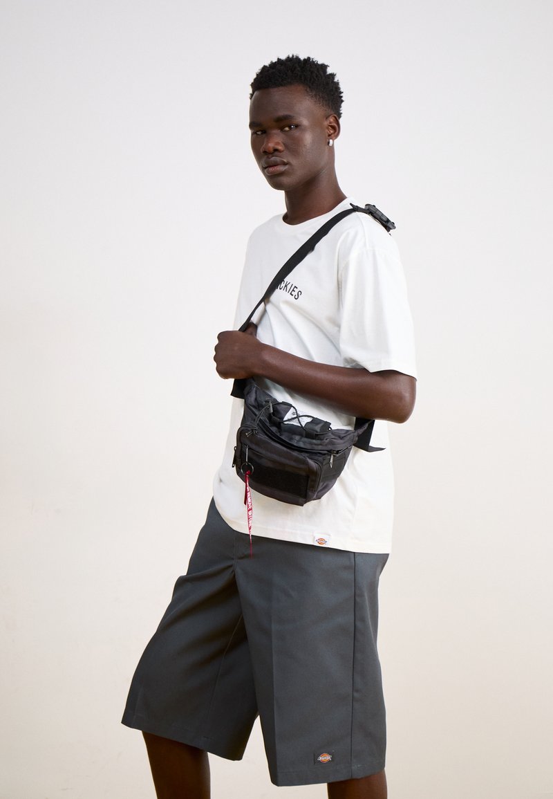Borsa a tracolla nera con più scomparti con zip e anello per portachiavi. Indossata con una t-shirt bianca e pantaloni corti grigi, con un logo.