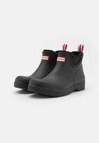 Bottes de cheville en caoutchouc noir avec des semelles texturées, comportant des languettes de traction rouges et blanches et une étiquette rectangulaire portant le texte "HUNTER".