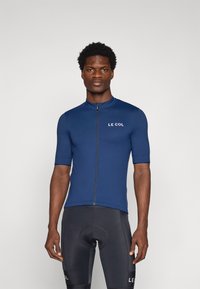 Maillot de cyclisme bleu marine à manches courtes, col haut et fermeture éclair à l'avant. Présente le logo "LE COL" en blanc sur la poitrine. Porté avec un short de cyclisme noir.