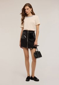 Top beige a maniche corte con bordi neri, abbinato a una mini gonna nera con dettagli lucidi sulle tasche e una cerniera fronte. Borsa e scarpe nere.