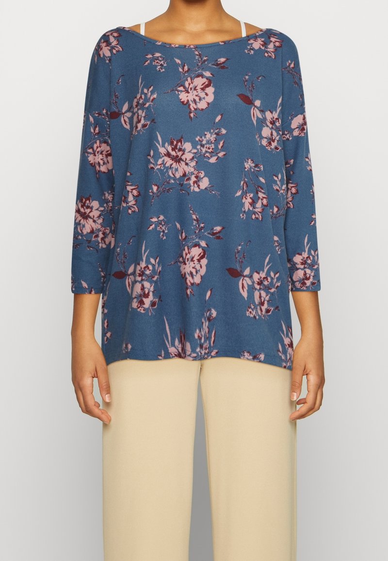 Blåaktig topp med trekvartsärmar, rosa blommönster, rundhalsning och en avslappnad passform, i kombination med ljusbeiga byxor.