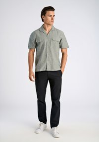 Camisa de manga corta con cuello en tejido verde claro, con dos bolsillos en el pecho, combinada con pantalones negros y zapatillas blancas sobre un fondo neutro.
