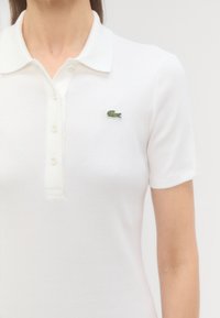 Biely rebrovaný polo tričko s krátkymi rukávmi, so zeleným vyšitým logom krokodíla na ľavej hrudi a gombíkovou légou.