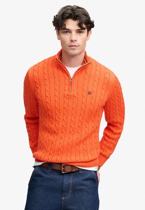 Ung mand med mørkt krøllet hår iført en lys orange kabelstrikket sweater med en kvart lynlås og blå jeans.