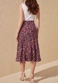 Nainen, jolla on pitkät aaltoilevat hiukset, pukeutuneena valkoiseen hihattomaan puseroon, korkeavyötäröiseen kuviolliseen midihameeseen ja valkoisiin korkokenkiin, seisoo sisätiloissa.