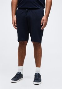 Άνδρας που φορά μπλε σκούρες βερμούδες με κορδόνι, λευκές κάλτσες και μπλε σκούρα αθλητικά παπούτσια, στέκεται σε απλό λευκό φόντο.