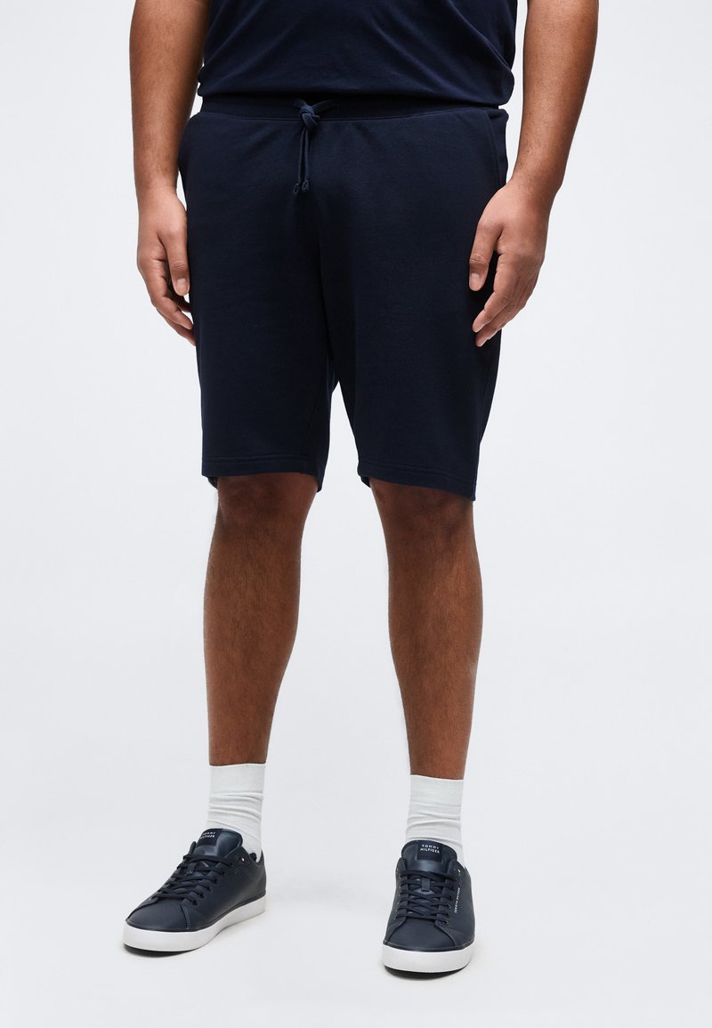 Άνδρας που φορά μπλε σκούρες βερμούδες με κορδόνι, λευκές κάλτσες και μπλε σκούρα αθλητικά παπούτσια, στέκεται σε απλό λευκό φόντο.