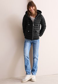 Doudoune noire avec capuche, présentant des coutures horizontales, une fermeture éclair et des poches avant. Associée à un jean bleu clair et des baskets blanches.