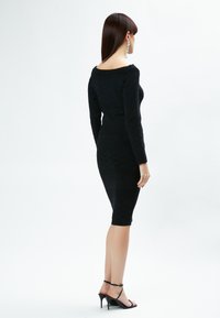 Figurbetontes schwarzes Strickkleid mit langen Ärmeln, schulterfreiem Ausschnitt und strukturierten Mustern. Kombiniert mit schwarzen, Riemchen-hohen Absätzen.