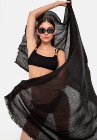 Svart lett skjerf med frynser, drapert dramatisk. Båret med en teksturert svart crop top. Modellen poserer med armene hevet, iført solbriller.