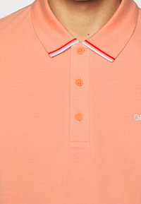 Vaalean oranssi pikeepaita, jossa on kaulus ja punavalkoinen raidoitus. Paidassa on neljä yhteensopivaa nappia ja pieni brodeerattu logo.