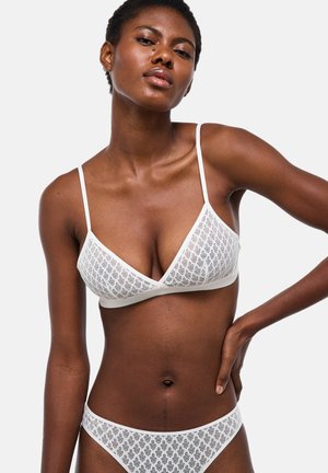 Femme portant un bralette en dentelle blanche et une lingerie assortie en dentelle, posant avec une main sur la hanche sur un fond clair uniforme.