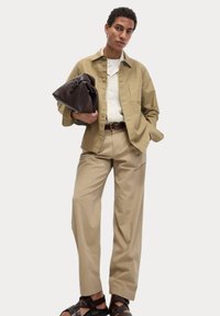 Een tan kleurig knoopshirt over een textuur wit sweater, gecombineerd met licht tan wijde broek en bruine sandalen met straps. Houdt een donkerbruine tas vast.