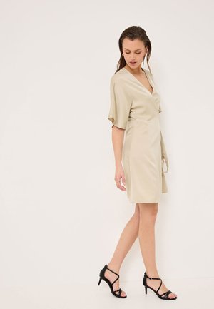 Abito avvolgente corto beige con maniche corte e scollatura a V, abbinato a tacchi neri con cinturini. Tessuto liscio con una leggera lucentezza.