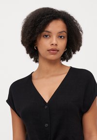 Blusa nera a maniche corte con scollatura a V profonda e tre bottoni frontali. Il tessuto presenta una finitura testurizzata. Design semplice e minimal.