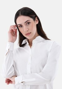 White, long-sleeve button-up shirt with a classic collar and cuff details, smooth texture, and subtle sheen. Model touches her hair.
