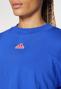Royal blue cotton t-shirt with a crew neck, featuring a red and white Adidas logo stitched on the chest. Smooth texture.
