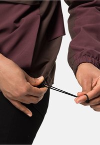 Veste bordeaux et marron avec des poignets élastiques ; une main ajuste le cordon de serrage à la taille, mettant en valeur la texture lisse du tissu et ses superpositions.