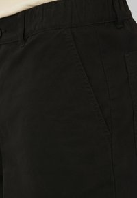 Gros plan sur la zone de la poche d'un pantalon noir avec des coutures visibles, une ceinture, un bouton, et une partie d'un haut beige rentrée à l'intérieur.