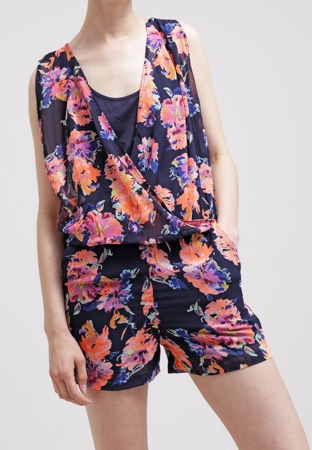 Femme portant une combinaison sans manches bleu marine à imprimé floral rose et orange, avec un col drapé et des poches latérales.
