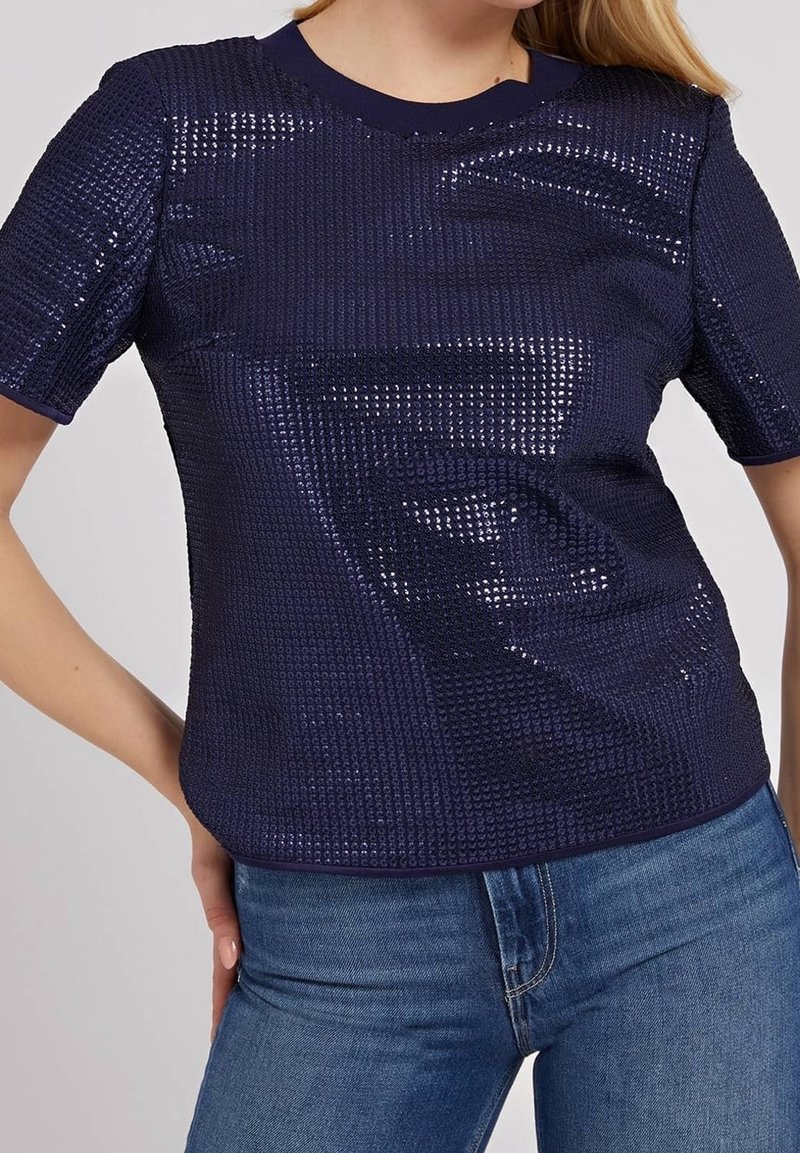 Femme portant un haut court bleu marine près du corps avec des paillettes, associé à un jean bleu, sur un fond uni.