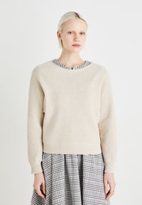 Beige kötött pulóver hosszú ujjakkal és bordázott szegéllyel, amit egy szürke és fehér kockás, bővülő fazonú szoknyával párosítanak.