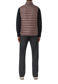 Gilet matelassé marron clair avec des coutures horizontales, un col montant, sans fermetures apparentes, porté avec un jean foncé et des chaussures claires.