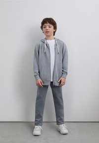 Sweat à capuche gris zippé avec cordons, associé à un T-shirt blanc et un pantalon gris. Chaussures de sport blanches. Design de tenue simple et décontractée.