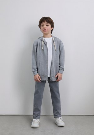 Sudadera con cremallera gris y cordones, combinada con una camiseta blanca y pantalones grises. Zapatillas deportivas blancas. Diseño de atuendo simple y casual.