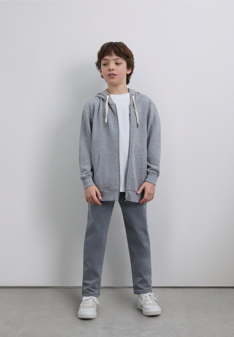 Sweat à capuche gris zippé avec cordons, associé à un T-shirt blanc et un pantalon gris. Chaussures de sport blanches. Design de tenue simple et décontractée.