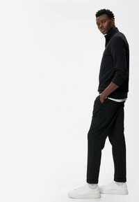 Pull en col roulé noir avec texture côtelée, associé à un pantalon noir tapered et des baskets blanches, avec des poignets retroussés et un design sobre.