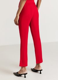 Red tailored trousers with a smooth, lightweight fabric and a slim fit, featuring a waistline and subtle back pocket detail. Black pointed heels with silver accents.