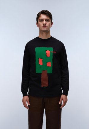 Man wearing black long-sleeve shirt with a green and brown tree graphic and orange abstract shapes, standing against a plain light background.