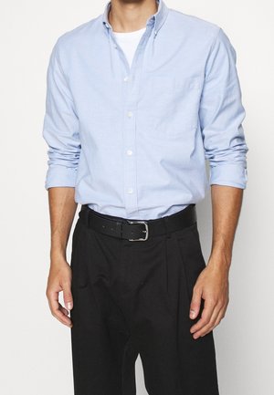 Mężczyzna noszący jasnoniebieską koszulę zapinaną na guziki na białą koszulkę, czarny pasek i czarne spodnie z podwiniętymi rękawami do przedramion.