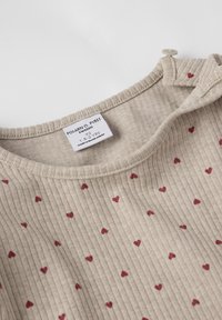 Beige ribbet top med små maroon hjertepatterner, med en knapdetalje ved skulderen og et mærke, der angiver mærke og størrelse.