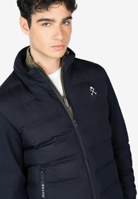 Chaqueta acolchada navy con cuello alto, cierre con cremallera y bolsillo frontal. Presenta un tejido acolchado y un pequeño logo en el pecho.