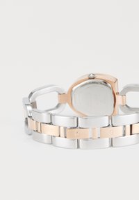 Ceas din metal argintiu și aur roz, cu un design voluminos al brățării. Prezintă accente cu logo gravat și un finisaj lucios pe carcasă și linkuri.