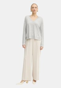 Donna con un maglione grigio chiaro a V, pantaloni larghi crema e tacchi bianchi con tacchi a cerchio dorati, in piedi su uno sfondo bianco.