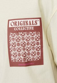 Sweatshirt beige clair présentant un graphisme bordeaux avec le texte "ORIGINALS COLLECTIVE" et un motif géométrique de soleils et de triangles.