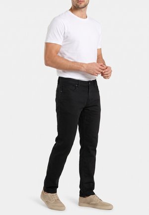 Hombre con una camisa blanca de manga corta lisa, vaqueros negros y zapatillas beige, de pie con las manos entrelazadas delante.