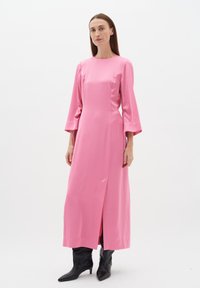 Rochie lungă din satin roz, cu gât rotund și mâneci evazate; are o deschidere frontală și este asortată cu cizme negre până la genunchi.