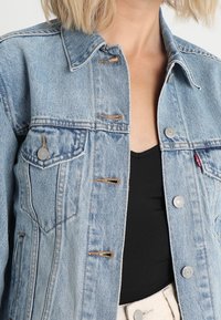 Světlá modrá džínová bunda s klasickým límcem, zapínáním na knoflíky, náprsními kapsami a mírně opotřebovaným vzhledem.