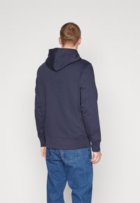Felpa con cappuccio blu navy in cotone misto, dotata di tasca a marsupio e polsini a costine, mostrata da dietro con jeans in denim a vestibilità rilassata.